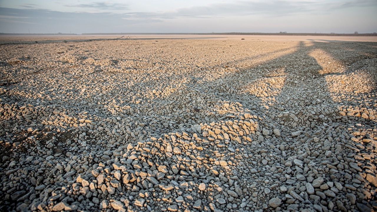 A wide, flat expanse of gravel and crushed stone under a hazy evening sky. Long shadows from something outside the frame, possibly tall structures or poles, stretch